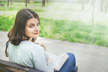 Obraz premium beautiful young girl with a book sits on a bench in the park and looks back at the camera. Back view.