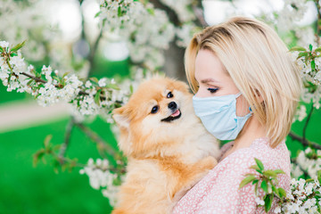 Young woman and red spitz with a medical mask on her face on nature on a spring day. Coronavirus pandemic