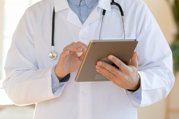 Close up of male doctor in white medical uniform and stethoscope consult hospital patient online using modern tablet gadget, man GP make notes of client anamnesis or history on Pad device
