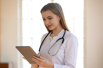 Young female doctor wearing white medical uniform and stethoscope busy working on modern tablet gadget, Caucasian woman GP typing texting consult patient online on Pad device, healthcare concept - obrazy, fototapety, plakaty