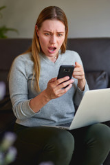 Sad woman sitting on sofa with laptop and talking on phone at home. College student studying on laptop and using phone.