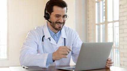 Smiling young male doctor in white medical uniform and wireless headset talk on video call on computer, consult patient online, happy Caucasian man GP in headphones watch webinar on laptop in hospital