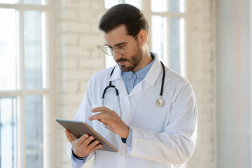 Serious young Caucasian male doctor in medical uniform and stethoscope fill patient anamnesis or history on modern tablet, man GP or physician consult client online using pad, healthcare concept - obrazy, fototapety, plakaty