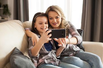 Obraz premium Beautiful young mother and her daughter are using a smartphone and smiling sitting on the couch in living room.