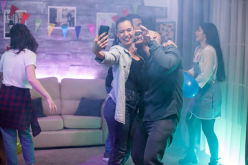Happy young man and woman taking a selfie at a party with their friends. Group of people dancing.