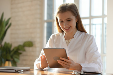 Smiling young female doctor wearing white medical uniform consulting patient online on tablet,...