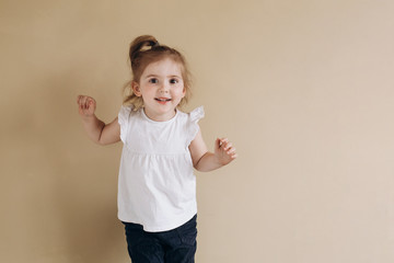 A happy little girl of European appearance stands on a beige background. Three year old baby in a white t-shirt.