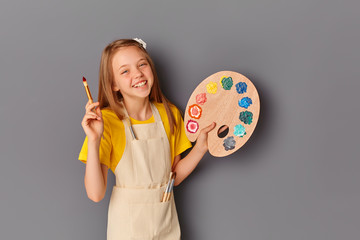 Portrait of a beautiful young teenage girl holding a wooden palette and brush against a gray wall. A smiling child with an art palette and an artist's brush in his hands.