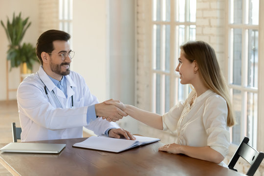 Smiling male Caucasian doctor in white medical uniform shake hand greeting getting acquainted with female patient at consultation, happy young man GP handshake client after closing deal in hospital