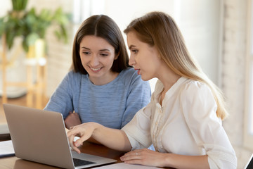 Smiling female employees sit at desk in office brainstorm discussing business project or plan on laptop together, happy woman colleagues cooperate work on modern computer gadget, teamwork concept