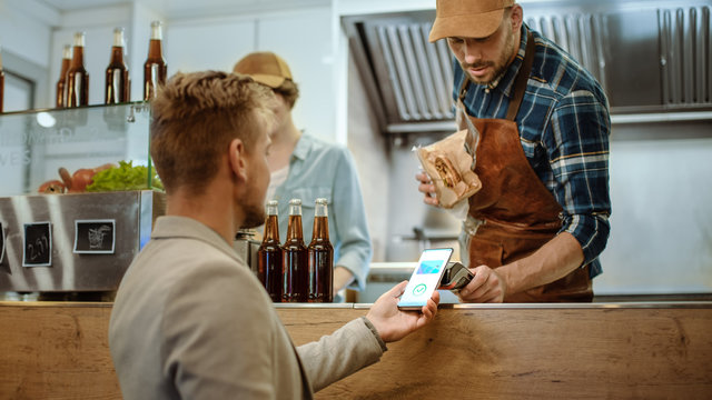 Food Truck Employee Hands Out A Freshly Made Burger To A Happy Young Male. Man In A Casual Suit Is Using His Smartphone For NFC Mobile Payment Solution. Street Food Truck Selling Burgers.