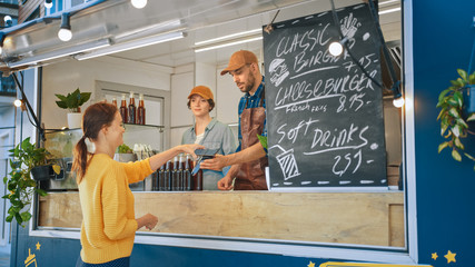 Food Truck Employee Hands Out a Freshly Made Burger to a Happy Young Female. Young Lady is Using NFC Mobile Payment to Pay for Food. Street Food Truck Selling Burgers in a Modern Hip Neighbourhood.