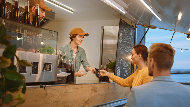 Food Truck Employee Hands Out A Freshly Made Burger To A Happy Young Female. Young Lady Is Paying For Food With Contactless Credit Card. Street Food Truck Selling Burgers In A Modern Hip Neighbourhood