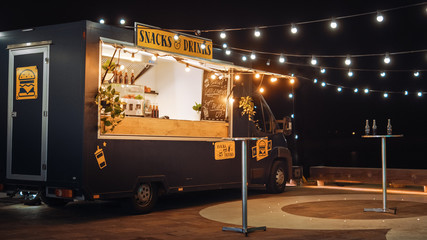 Empty Scene with a Dark Street Food Van Standing in the Evening in a Nice Warmly Lit Neighbourhood Next to the Sea. Food Truck Has Burgers and Drinks for Sale. Tables Have Bottles on Them. - Powered by Adobe