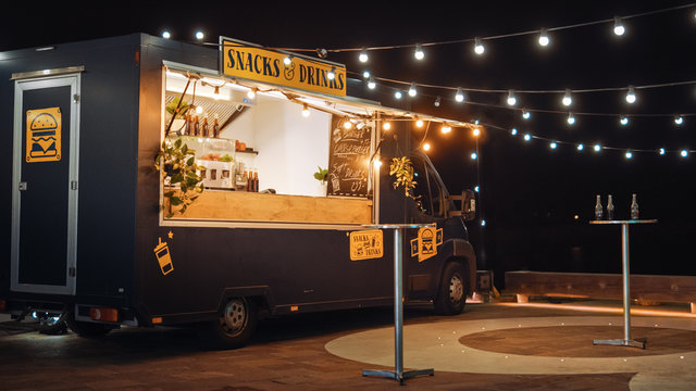 Empty Scene with a Dark Street Food Van Standing in the Evening in a Nice Warmly Lit Neighbourhood Next to the Sea. Food Truck Has Burgers and Drinks for Sale. Tables Have Bottles on Them.