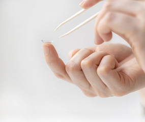 A hand with tweezers reaches for the contact lens on the forefinger of the other hand. Using contact lenses to correct vision.