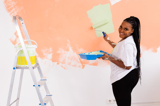 Beautiful african american girl painting the wall with paint roller. Portrait of a young beautiful woman painting wall in her new apartment. Redecoration and renovation concept.