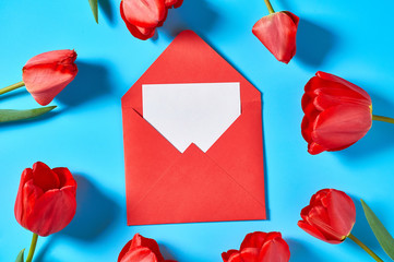 Scattered beautiful red tulips near envelope with blank paper sheet on blue background. Concept of celebrating valentine's, mother day or easter
