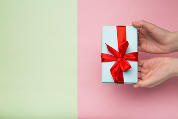 Top view of a woman's hands holding a white gift box with a red ribbon on a pink and green background. Flat lay. Gift for birthday, Christmas, Valentine's day, New year.  copy  space.