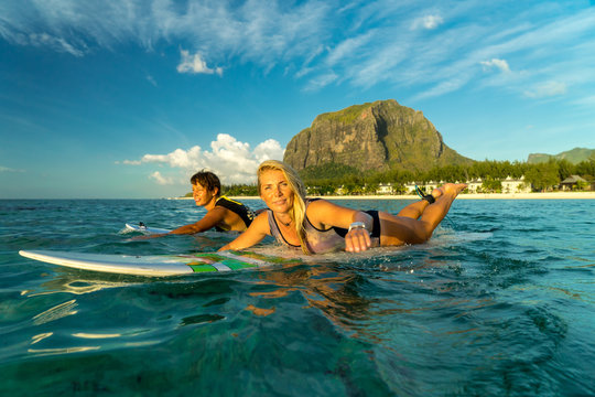 Two beautiful girls rowing on surfboards in the ocean against the backdrop of a picturesque mountain