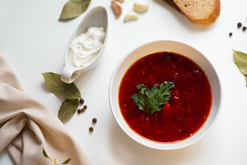 Beetroot soup in white bowl on a white table served with napkin, garlic, pepper, bread and saucy with sour cream. Traditional ukrainian, russian soup borscht with greens. Top view.