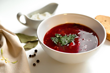 Beetroot soup in white bowl on a white table served with napkin, garlic, pepper, bread and saucy with sour cream. Traditional ukrainian, russian soup borscht with greens.