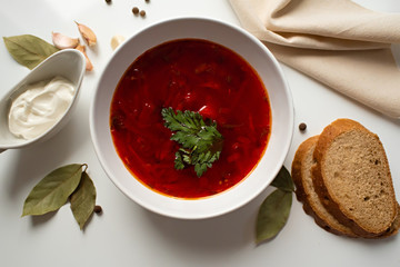 Beetroot soup in white bowl on a white table served with napkin, garlic, pepper, bread and saucy with sour cream. Traditional ukrainian, russian soup borscht with greens. Top view.
