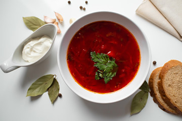 Beetroot soup in white bowl on a white table served with napkin, garlic, pepper, bread and saucy with sour cream. Traditional ukrainian, russian soup borscht with greens. Top view.