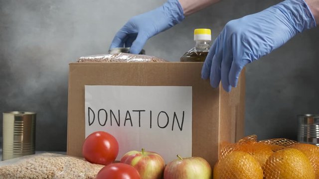 closeup on volunteer wearing blue protective gloves putting grocery food in carton donation box: vegetable oil, cereals, tin cans, tomatoes, fruits. Coronavirus relief funds, charity help, delivery