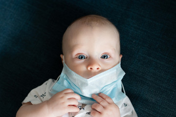 White caucasian baby boy playing with a face mask trying to take it of while staying at home on isolation and quarantine during pandemic of coronavirus virus covid-19 pulling mask down from the face