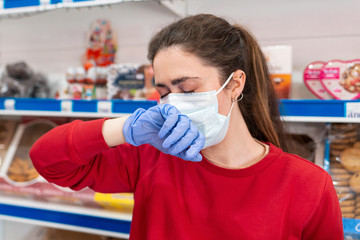 A woman in rubber gloves and a medical mask,suffering from a runny nose covering her face with her hand.In the background are shelves of the store.The concept of coronovirus and the crisis in business