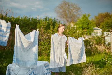 Blonde woman villager hanging wet white-blue laundry on clothesline to dry in the backyard. A green tree in the background.
