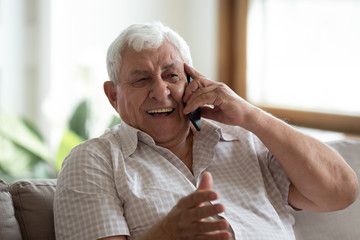 Overjoyed senior 80s grandfather sit on sofa at home laugh talking on modern cellphone, happy...