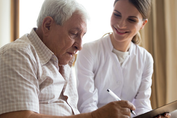 Obraz premium Smiling young female doctor or caregiver visit senior male client close deal at home, positive woman medical worker consult mature man sign insurance contract, offer help, elderly healthcare concept