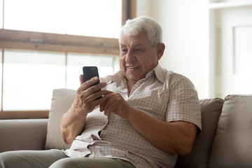 Smiling mature 80s man sit on sofa in living room texting messaging on modern smartphone gadget,...