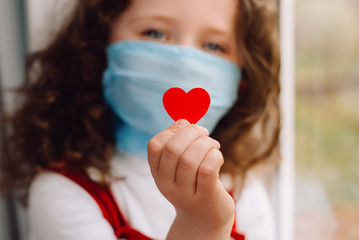 Cute preschool little girl in protective medical masks sitting on sill, holding red heart a way to show appreciation and to thank all essential employees during Covid-19 pandemics. Selective focus