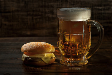 Cheeseburger with a glass of beer on a dark wooden table. Food on a black shabby background. Contrasting yellow light sparkles on the glass.
