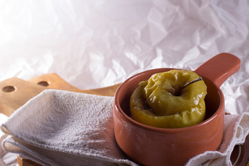 A baked apple in a clay pot on a linen cloth and wooden desk against white background