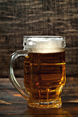 A glass of amber beer. Glasses on a wooden table. Black shabby boards on the background. Bright contrasting light and glare on the glass.