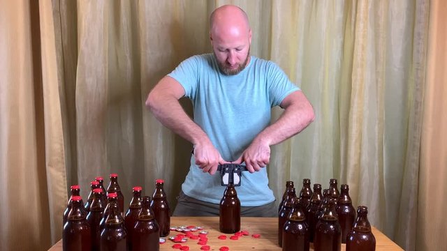 Craft beer brewing at home, man closes brown glass beer bottles with plastic capper on wooden table with red crown caps. Close up horizontal HD video.