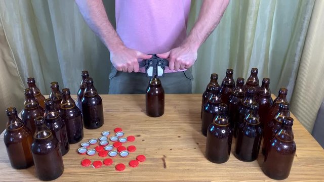 Craft beer brewing at home, man closes brown glass beer bottles with plastic capper on wooden table with red crown caps. Close up horizontal HD video.