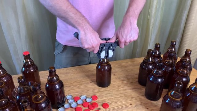 Craft beer brewing at home, man closes brown glass beer bottles with plastic capper on wooden table with red crown caps. Close up horizontal HD video.