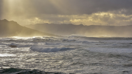 Big waves break on the coast, in the sunlit sea, passing through the clouds at sunset. Galicia, Spain.