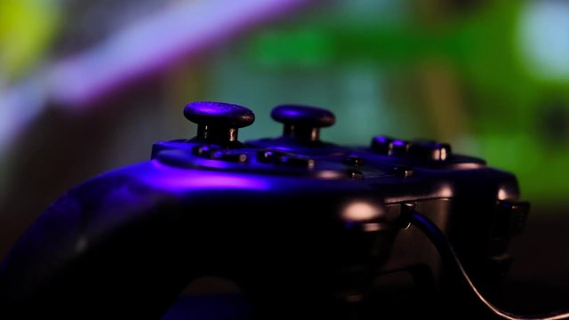 Black controller to play video games on game console. Gamepad in front of a large screen, projector or TV. Camera moves along table. Panoramic scene close-up view. Light changes to blue, red, green.