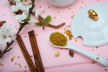 Bee pollen granules close up on ceramic white spoon. Bee raw product on pink wooden background, decorated with apricot blossom flowers and cinnamon sticks. Healthy supplement of apiery.