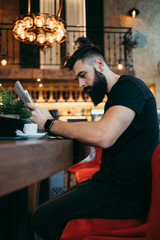 Fototapeta na wymiar Young adult hipster man with a long beard sitting in modern cafe bar, drinking coffee and reading newspapers.