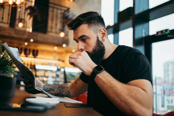 Young adult hipster man with a long beard sitting in modern cafe bar, drinking coffee and reading newspapers.