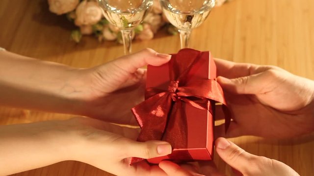 Man gives red present box with ribbon to woman. Indoors restaurant or home, romantic dinner. St. Valentine's Day, International Women's Day, birthday, holiday concept.