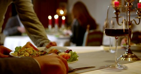 a waiter serves a restaurant dish on the table with a glass of wine and candles. male hands take a fork and knife. in the background you can see another couple out of focus