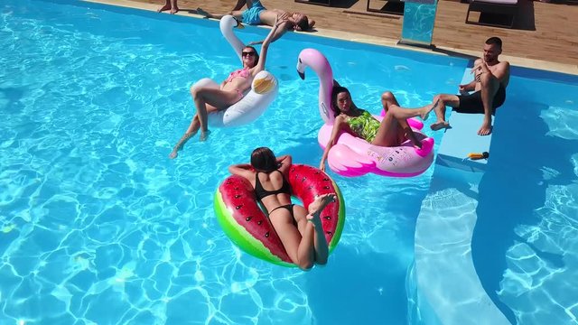 Aerial of friends having party in swimming pool with inflatable flamingo, swan, mattress. Happy young people wave hands at drone camera on luxury resort. View from above. Girls in bikini sunbathing.